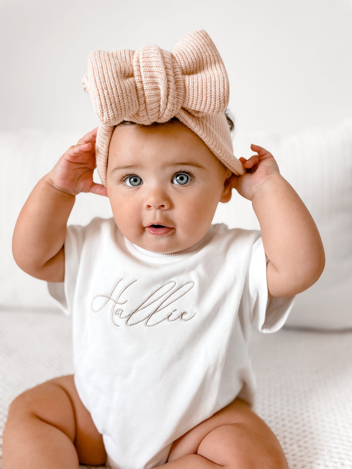 A baby wearing a white short-sleeve bodysuit with the name 'Hallie' in silver script and a large beige ribbed bow headband, sitting on a white textured surface.