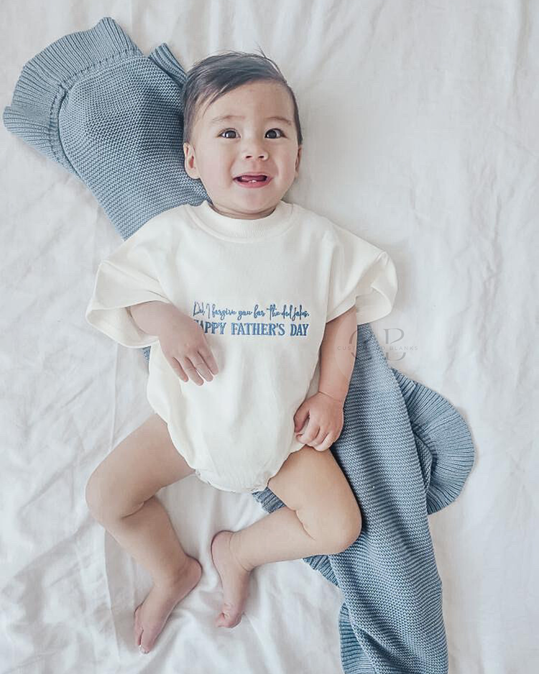 A baby lying on white bedding wearing a cream short-sleeve bodysuit with blue text that reads 'I'll remember you for Thanksgiving and Happy Father's Day.' A blue knit blanket is behind the baby's head, and a striped blue-and-white throw blanket is draped to the right.